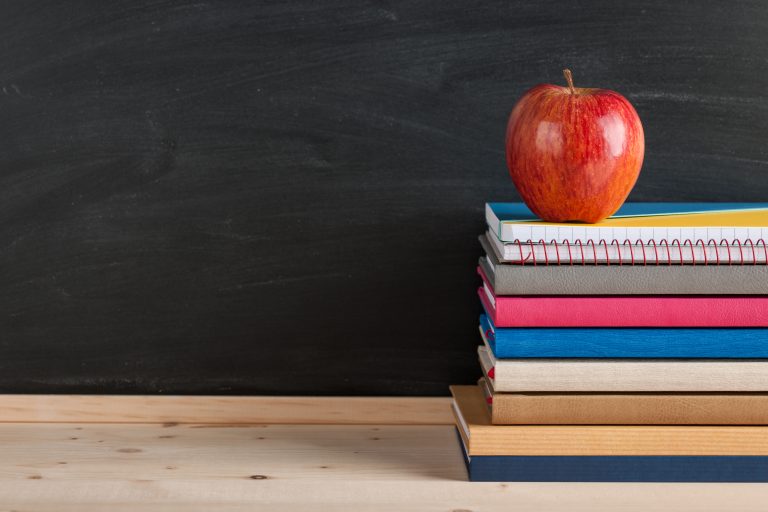 Wie bei einer Ausbildung liegen ein Stapel Bücher und ein Apfel auf einem Holztisch vor einer schwarzen Tafel.